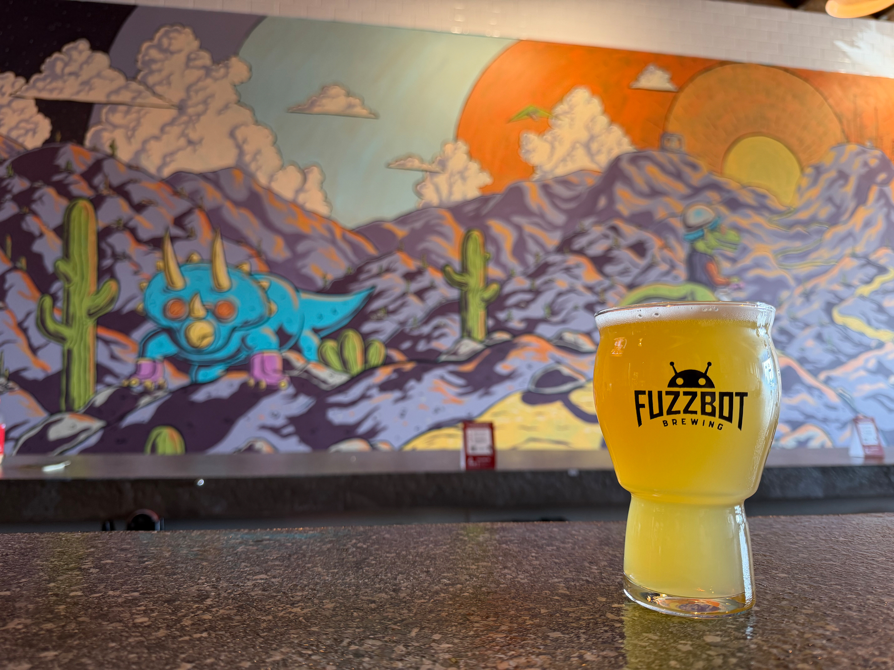 A glass of Fuzzbot Brewing beer sits on a counter in front of a colorful mural depicting a desert landscape with cacti and mountains.
