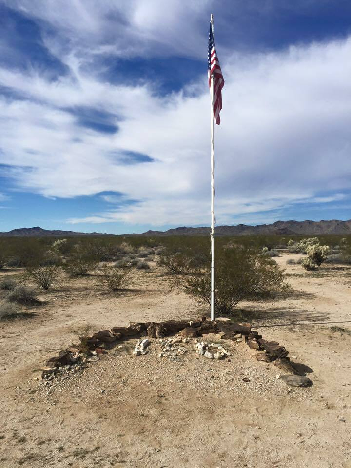 Auto-generated description: A flagpole with an American flag stands in a desert landscape with a ring of stones at its base.