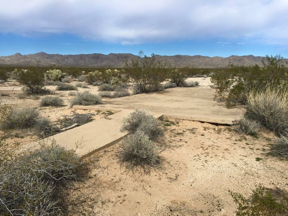 Auto-generated description: A flat, cracked concrete slab is situated in a desert landscape with sparse shrubs and mountains in the background.