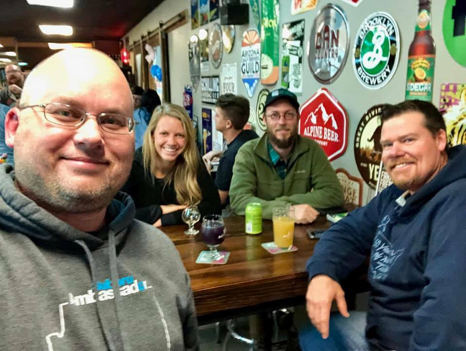 Auto-generated description: A group of four people is smiling at a table in a bar with various beer signs on the wall.