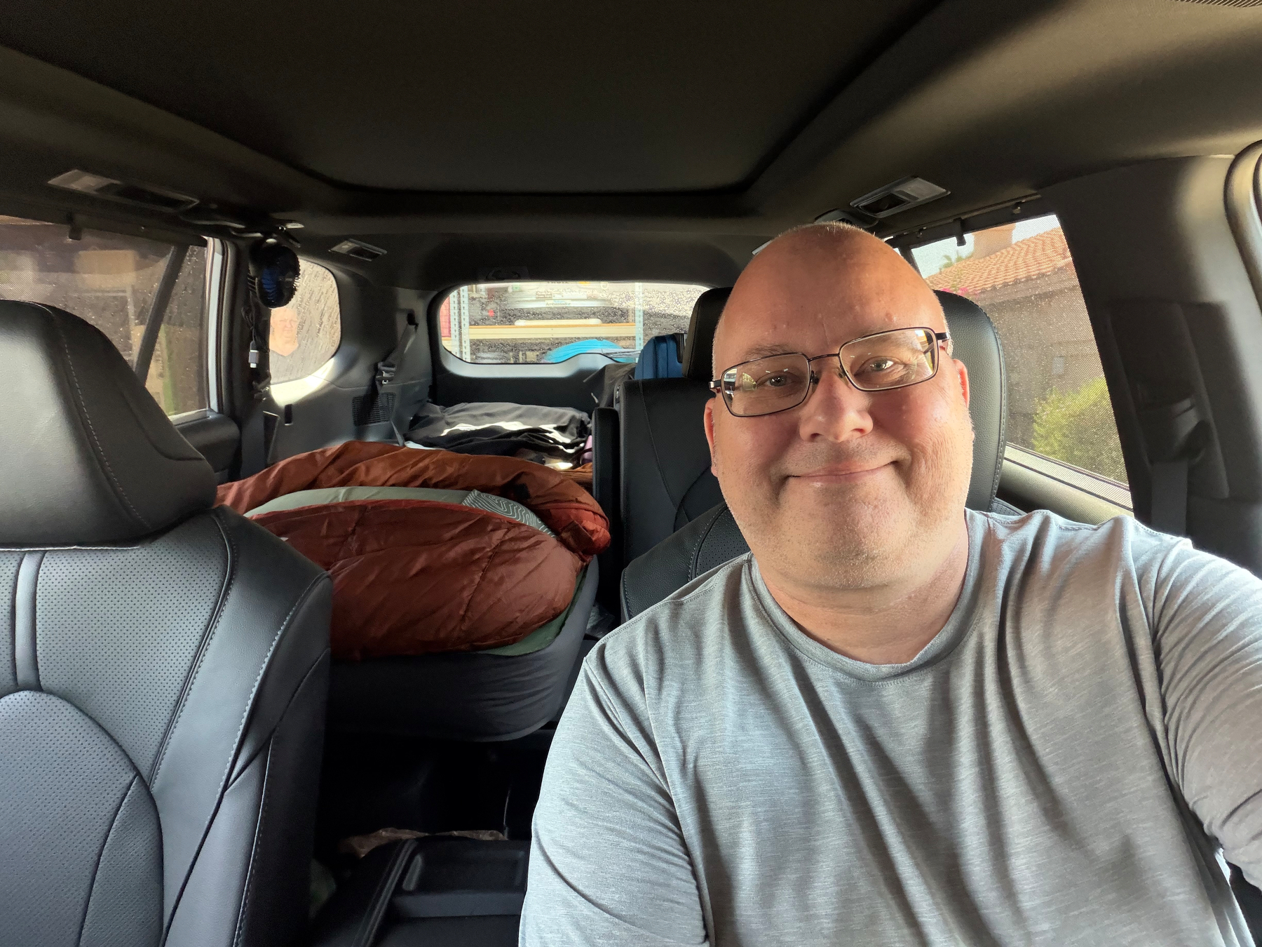 Auto-generated description: A smiling man wearing glasses and a gray shirt is sitting in the driver's seat of a vehicle with a sleeping bag and other gear in the back.