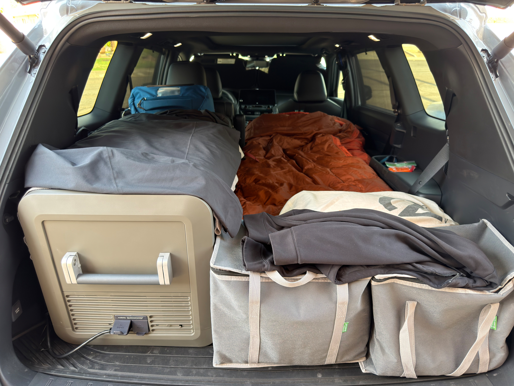 Auto-generated description: A car trunk is set up with packed storage bags, a portable refrigerator, and a sleeping bag, indicating preparation for camping or travel.